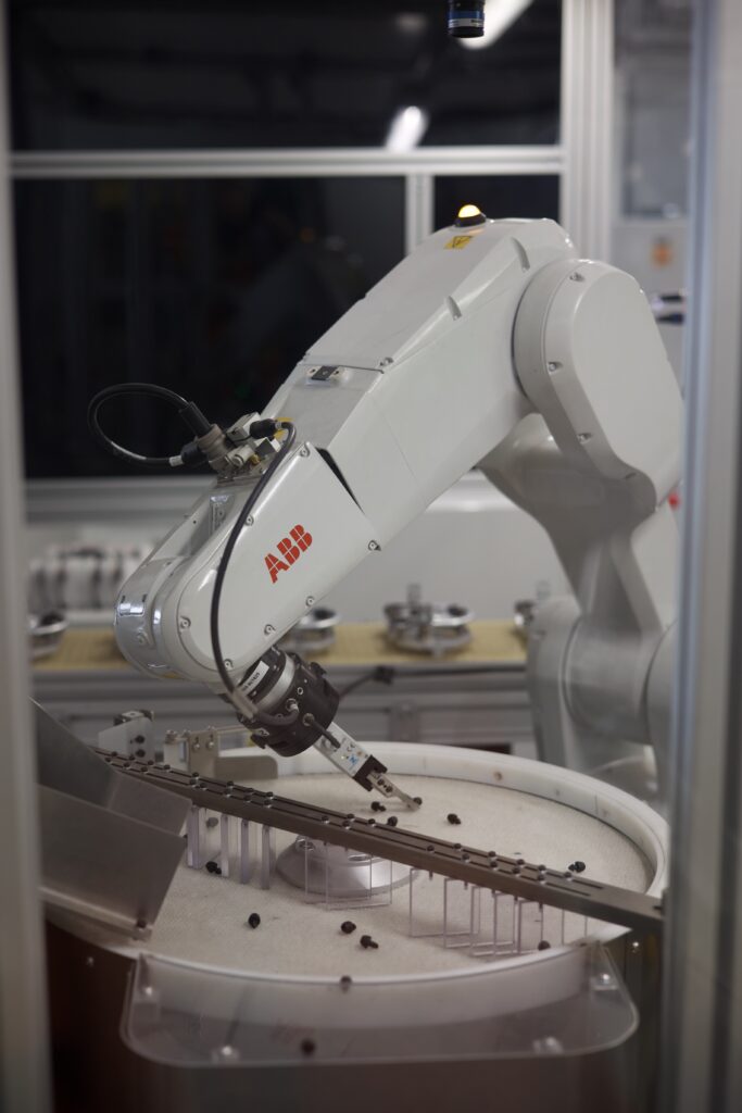 A white ABB industrial robotic arm equipped with a gripper is operating inside an automated manufacturing cell. The robot is picking small black components from a FlexiBowl flexible feeder, which has a rotating white disc and a vision-guided system. A conveyor system is positioned nearby to transport sorted parts. The background features a clean, high-tech production environment with additional machinery and structured lighting