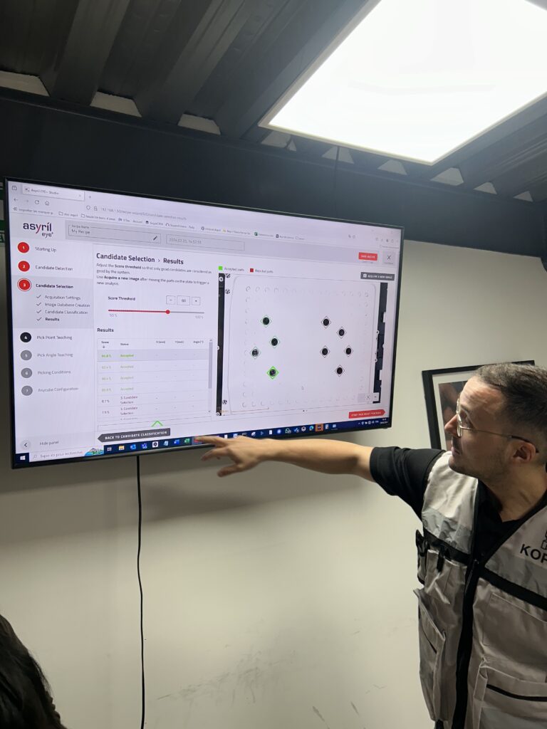 A technician in a branded vest points at a large screen displaying the Asyril Eye+ software interface, which shows candidate selection results for part detection in a flexible feeding system. The setup appears to be part of a training or testing session in an industrial or lab environment.
