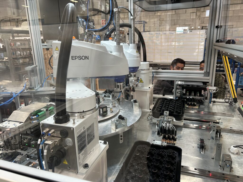 An automated assembly system featuring an Epson SCARA robot with a rotary indexing table and modular tray loading for part handling. The enclosed workstation includes sensors, actuators, and vision systems for precision assembly. Two individuals observe the machine, indicating a testing or calibration process in an industrial setting.